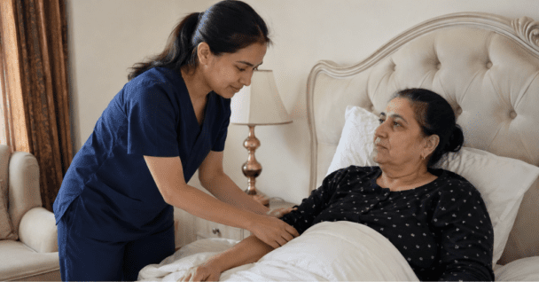 A professional home nurse in dark blue scrubs gently assisting an elderly female patient resting in bed in a home care setting.